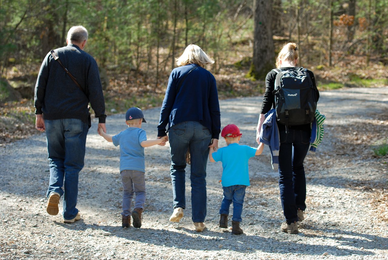 Two adults and two children walk hand in hand on a gravel trail surrounded by trees.