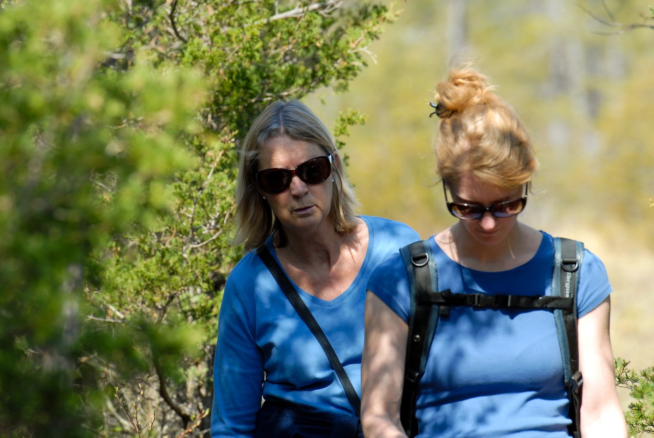 Two women wearing sunglasses and blue shirts walk along a wooded trail, one carrying a backpack.