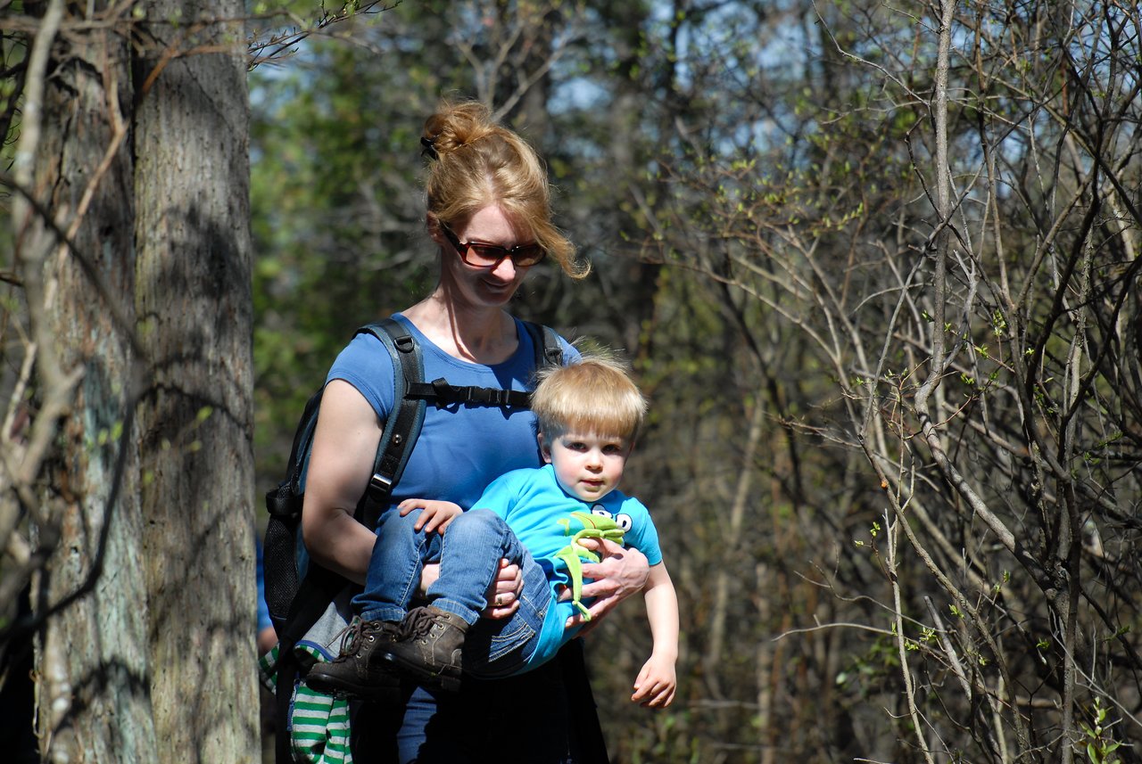A woman wearing a backpack carries a young child while walking on a wooded trail.