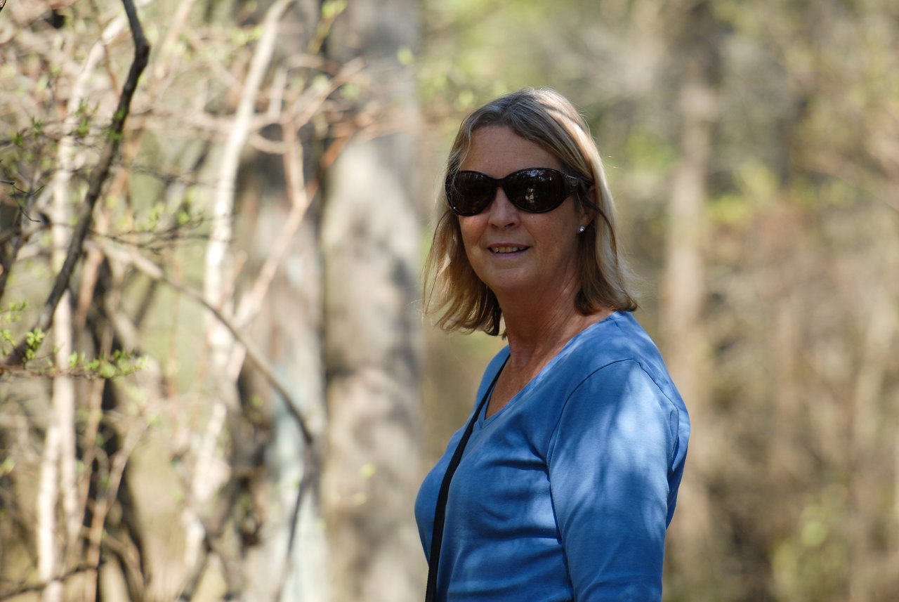 A woman wearing sunglasses and a blue shirt stands on a wooded trail, smiling at the camera.
