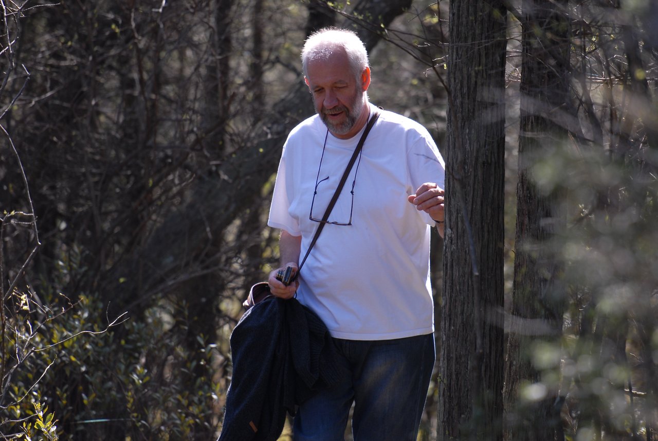 A man in a white shirt walks along a wooded trail, holding a jacket and a small object in his hand.