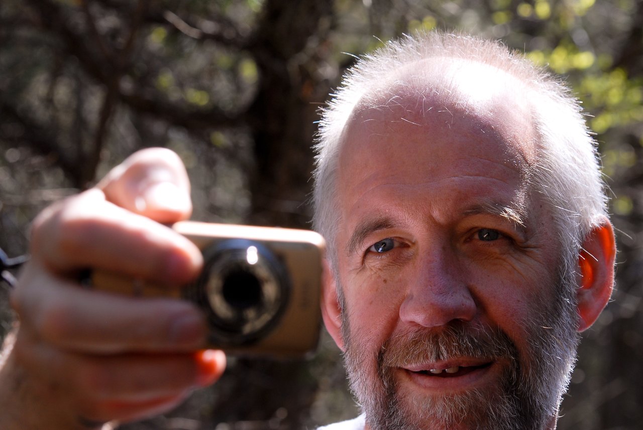 A man with a beard holds a small camera and takes a photo while standing on a wooded trail.