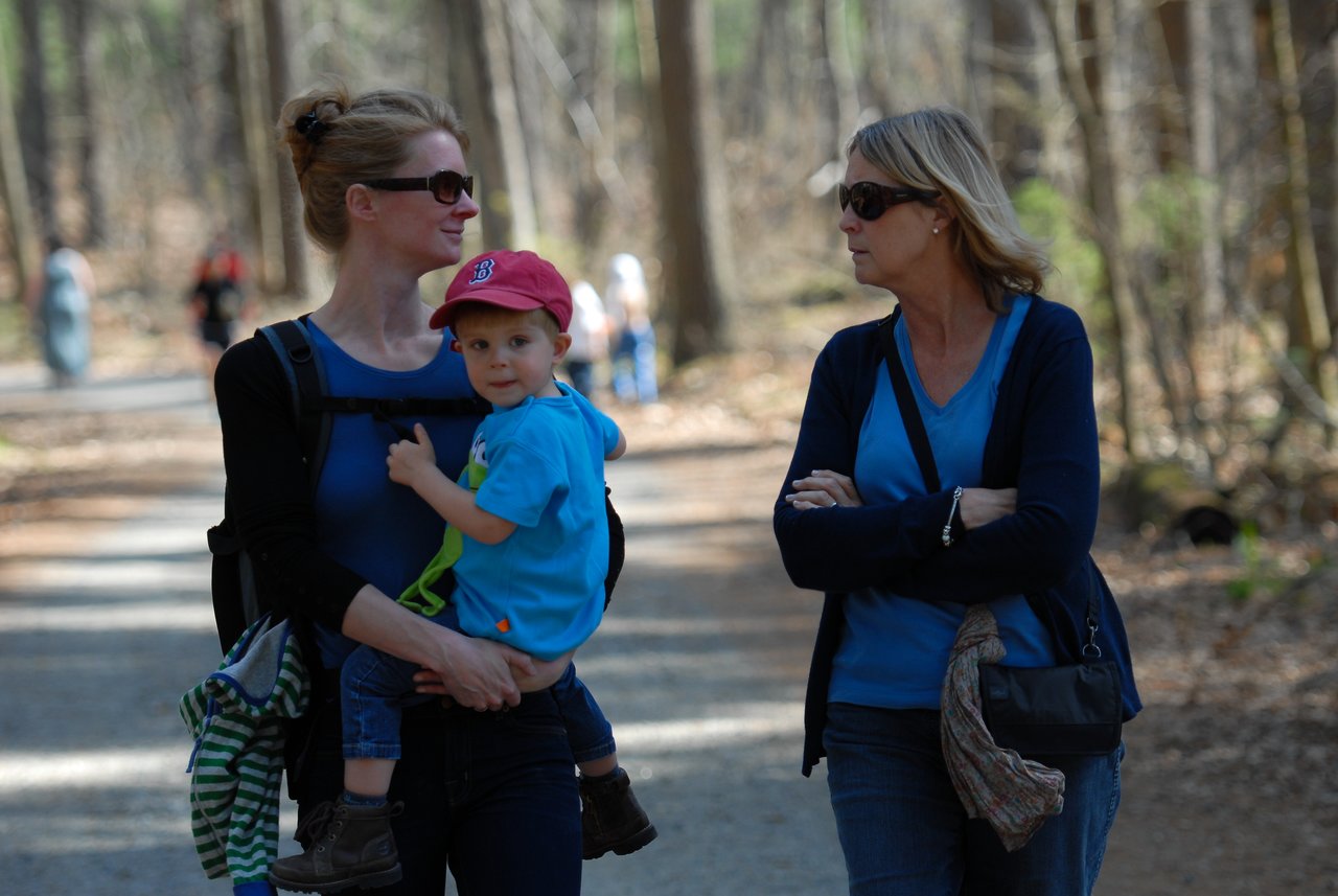 A woman carries a young child while talking to another woman on a wooded trail.