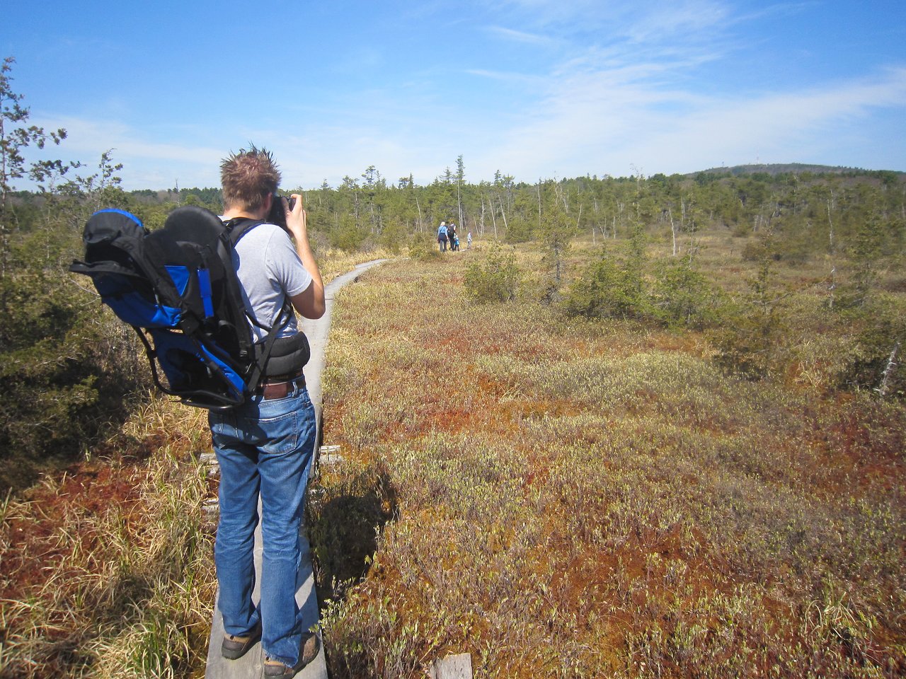 A person with a child carrier takes a photo on a narrow wooden trail, with others walking ahead.