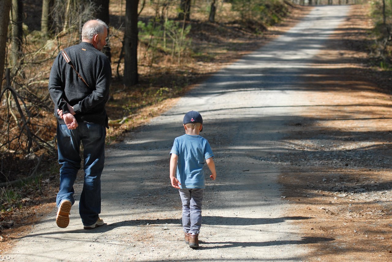 An older man and a young child walk along a dirt trail surrounded by trees.