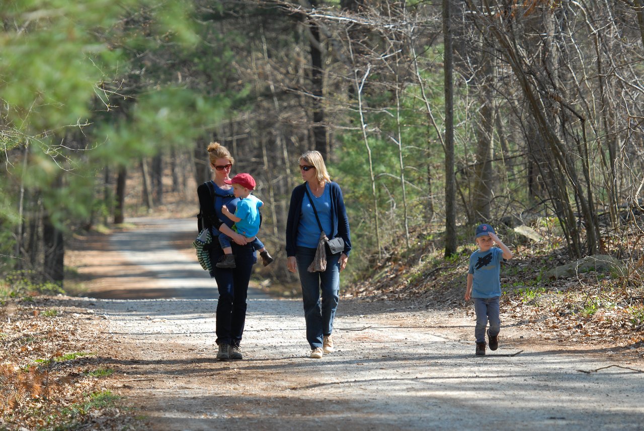 Two women walk along a forest trail, one carrying a young child, while an older child walks ahead.