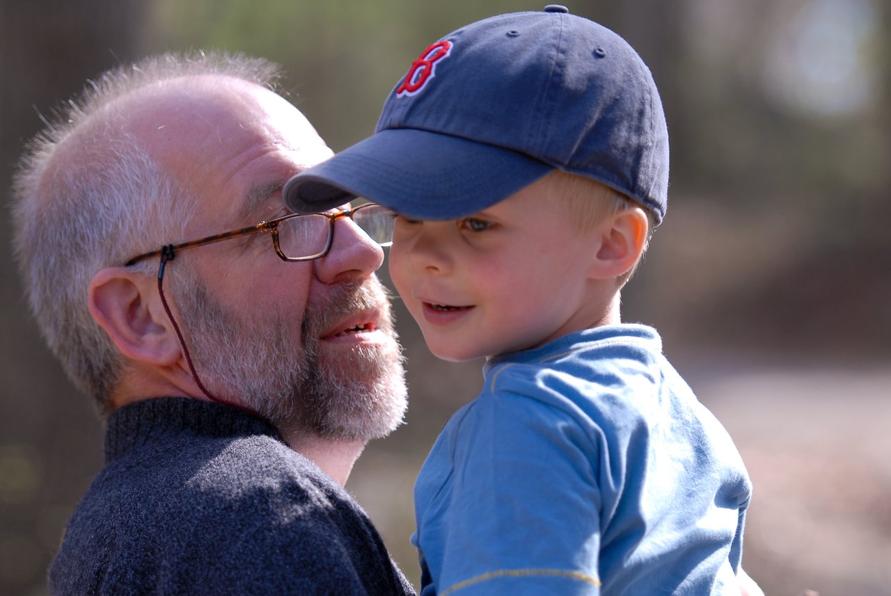 An older man with glasses holds a young boy wearing a blue cap and shirt on a wooded trail.