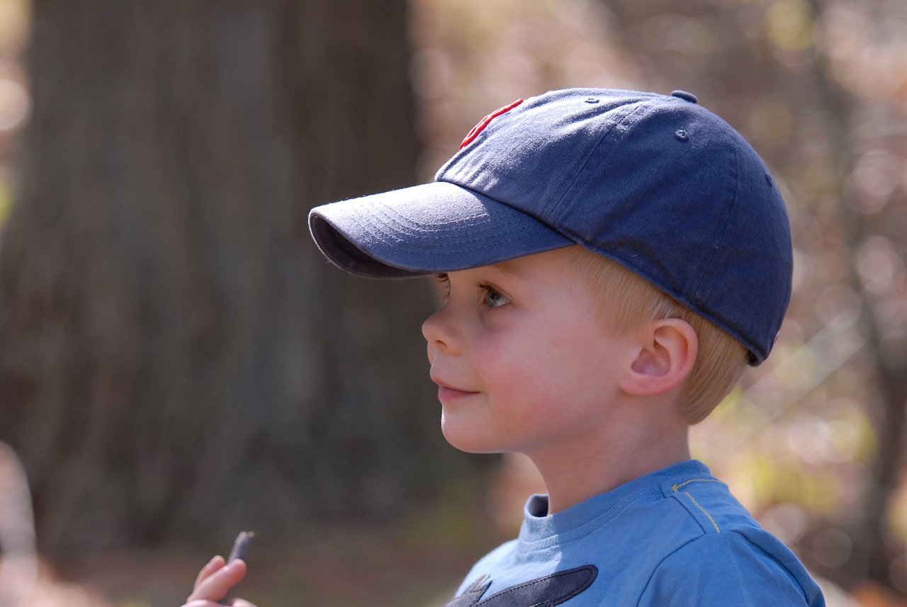 A young child in a blue cap and shirt looks to the side while holding a small object outdoors.