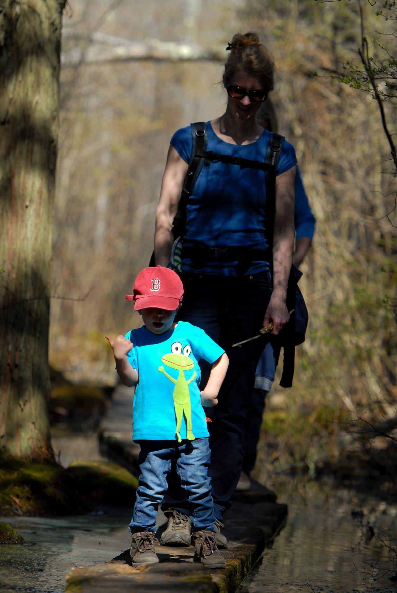 A child in a red cap walks on a narrow trail, followed closely by an adult wearing a backpack.