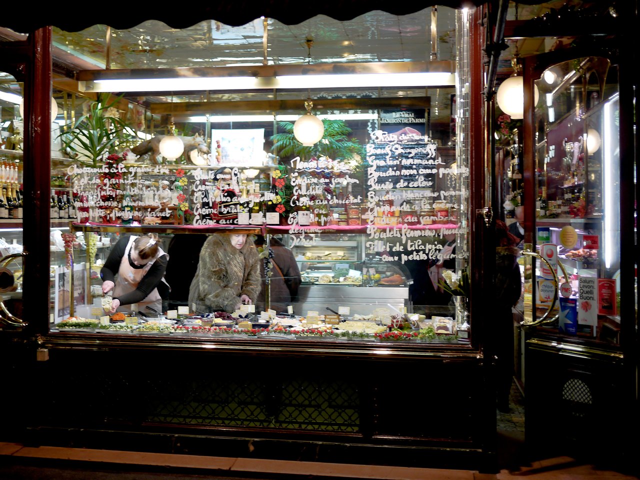 A woman in a fur coat shops at a butcher counter in Paris at night.