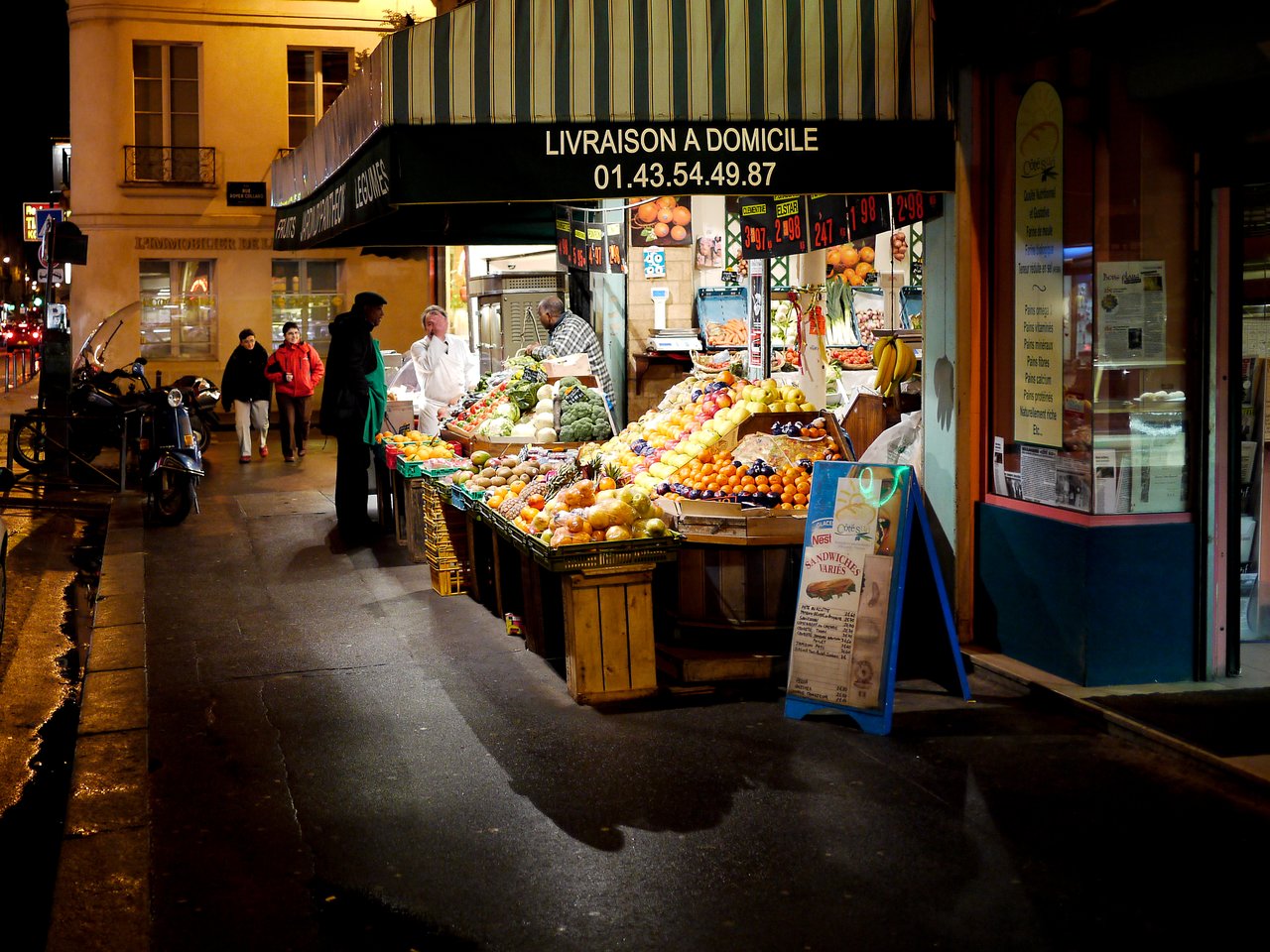 A fruit and vegetable stand at night with merchants and customers interacting on a well-lit Paris street.