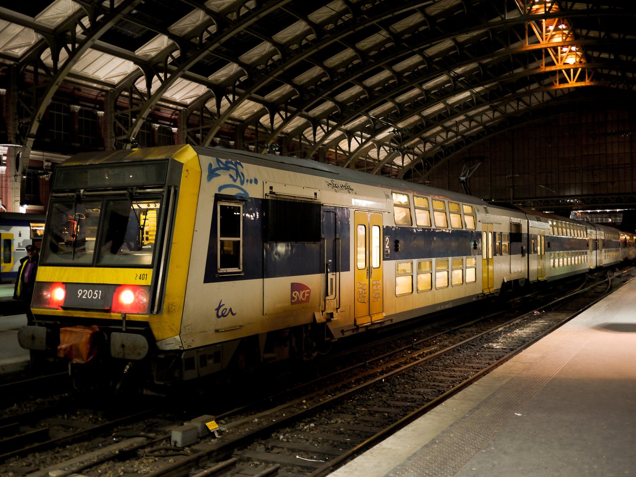 A double-decker SNCF train is stopped at a well-lit station platform at night, with passengers nearby.