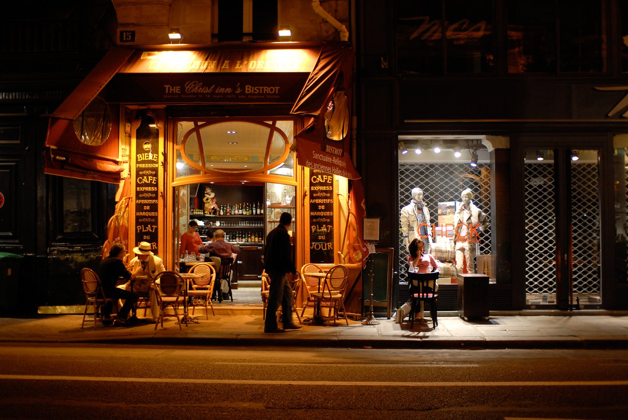 People sit at outdoor tables of a warmly lit bistro at night, while others dine inside.