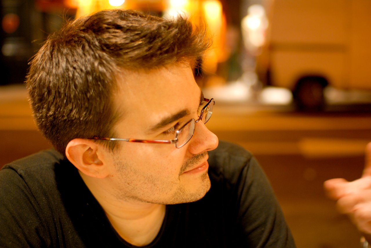 A man wearing glasses and a black shirt listens attentively while someone gestures in conversation.