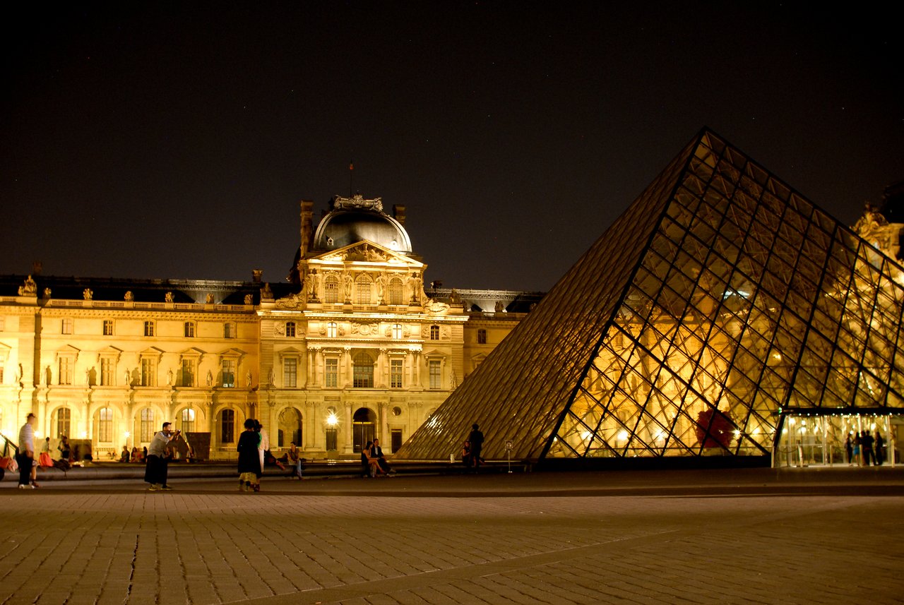 The Louvre Museum and its glass pyramid are illuminated at night, with people walking and sitting nearby.