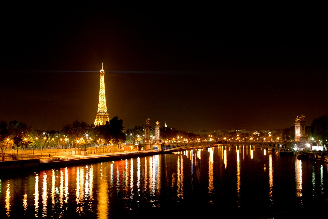 The Eiffel Tower glows at night, reflecting on the Seine River, with streetlights illuminating the surrounding area.