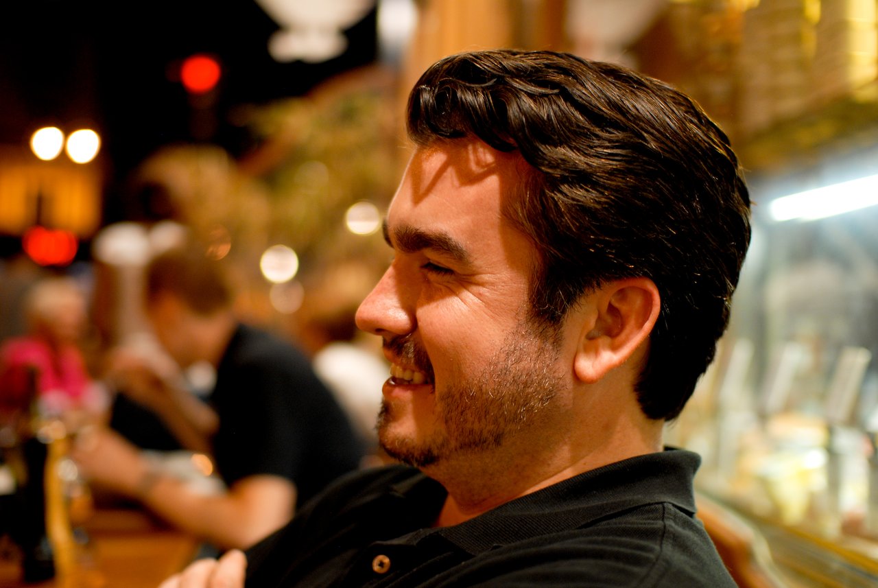 A man in a black shirt smiles while sitting in a warmly lit restaurant or café.
