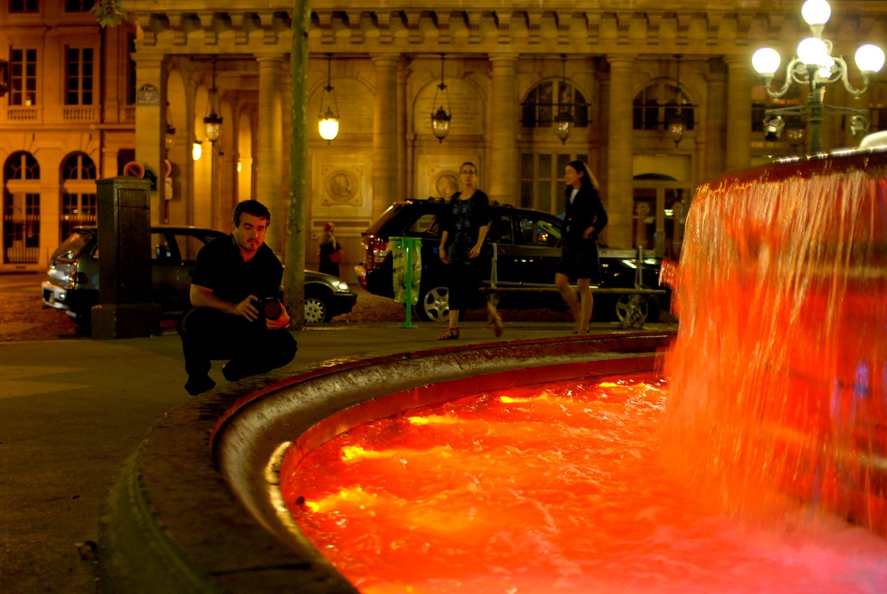 A man crouches near a fountain with red-lit water at night, holding a camera.