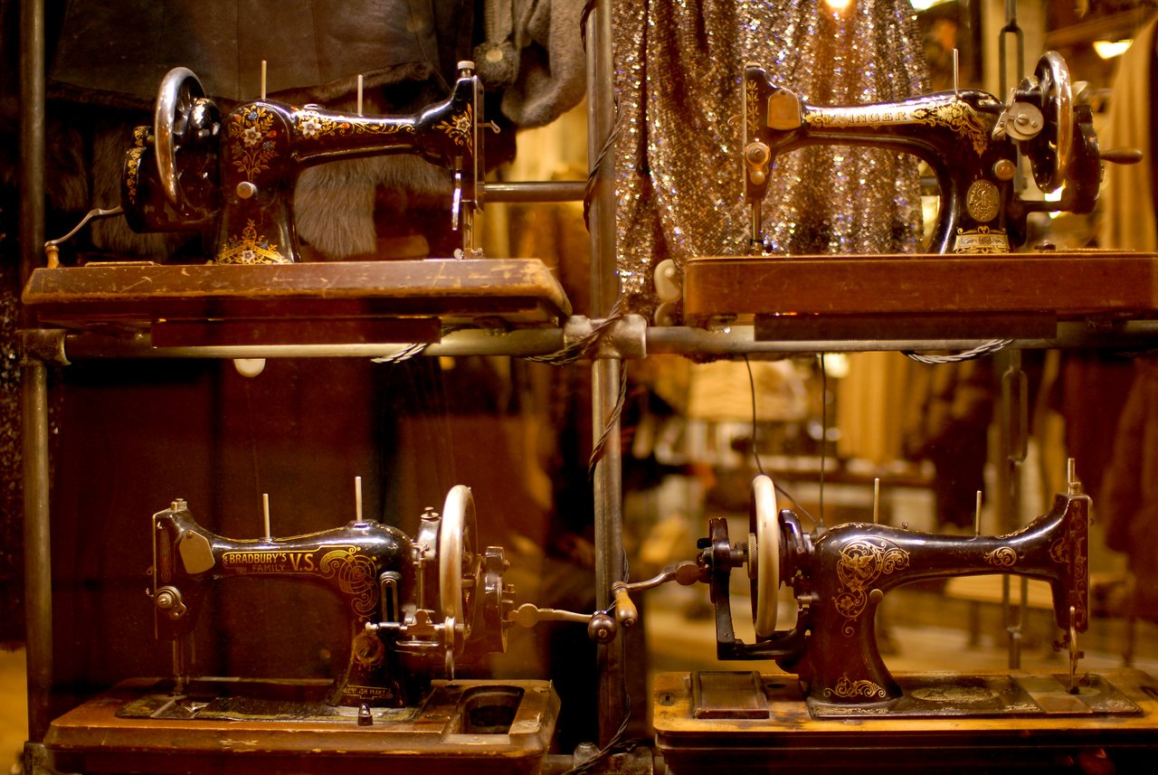 Four antique sewing machines displayed on metal shelves in a store window, with clothing in the background.