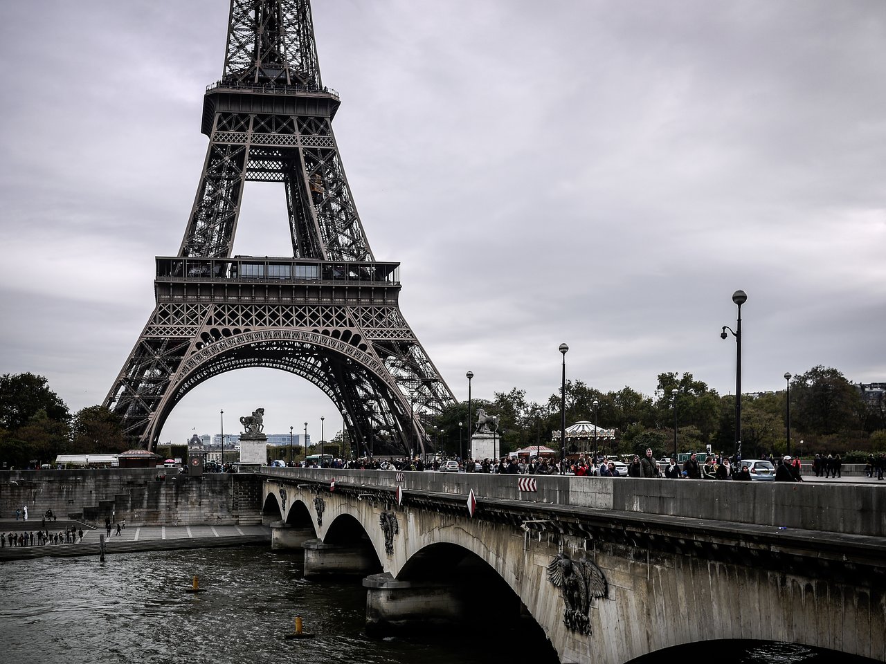 The Eiffel Tower stands tall behind a busy bridge with pedestrians, cars, and streetlights on a cloudy day.