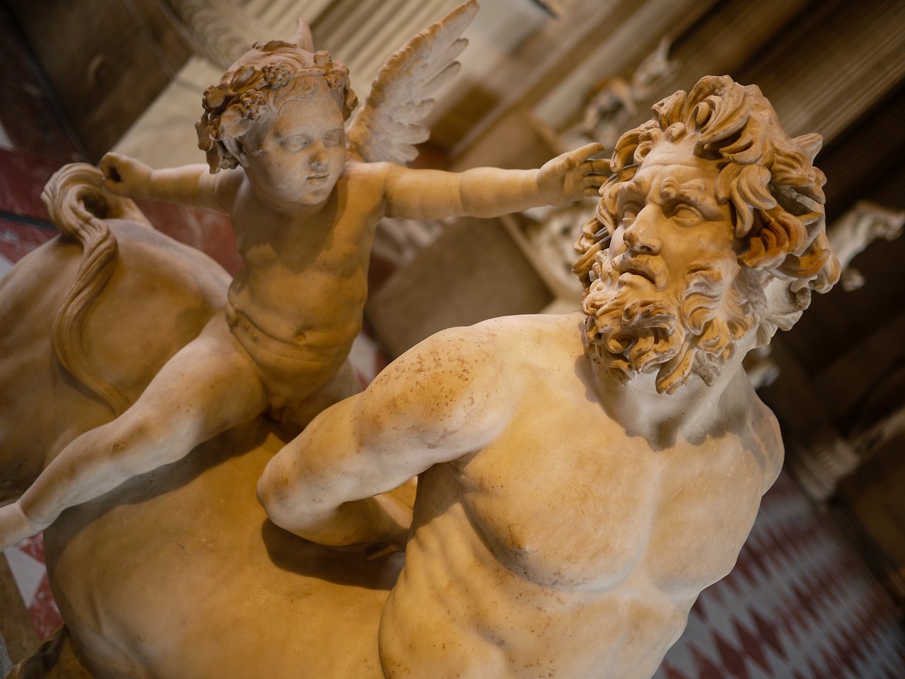 Marble statue of a bearded man with a muscular torso and a winged child reaching toward his head.