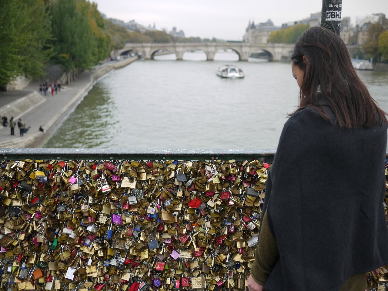 A woman stands by a bridge railing covered in love locks, looking at the river and passing boat.