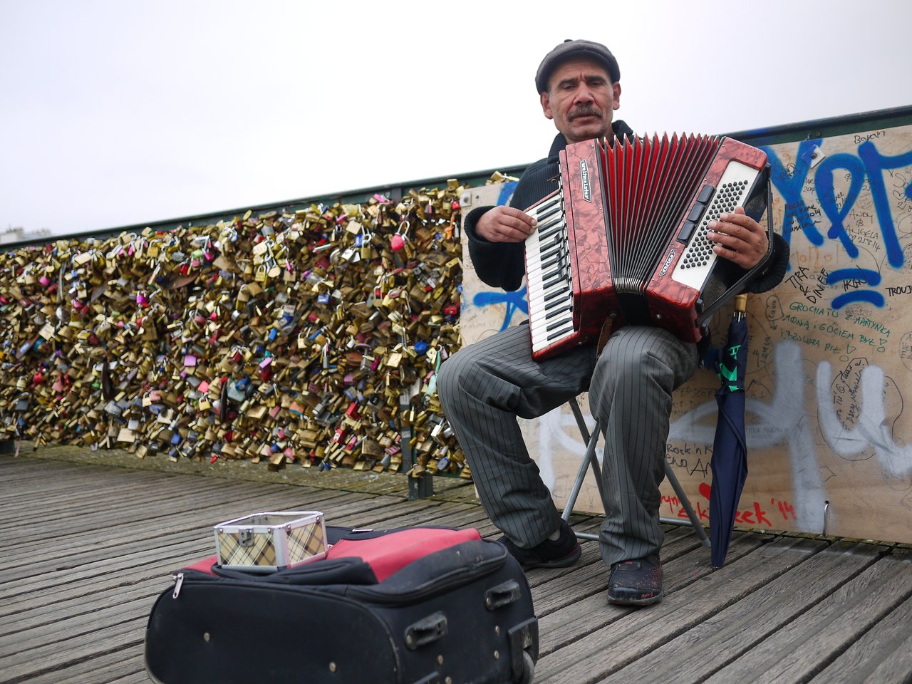 A street musician sits on a folding chair, playing an accordion near a fence covered in padlocks.
