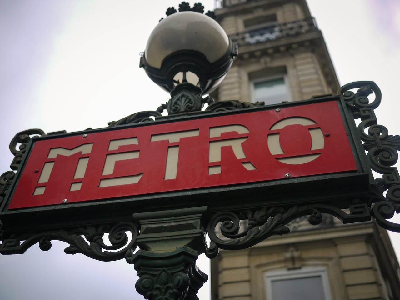 A red Paris Metro sign with decorative metalwork, mounted on a post, with a building in the background.