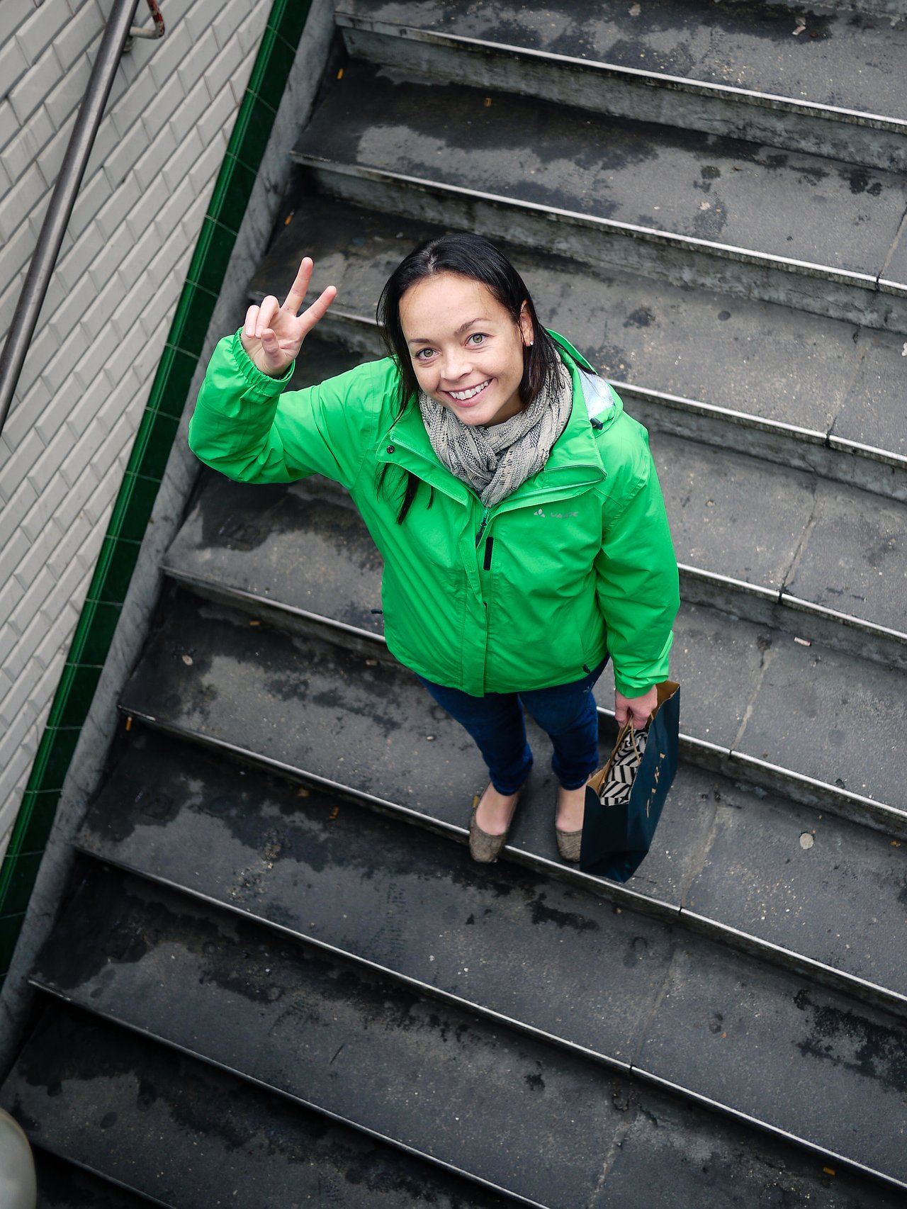 A woman in a green jacket stands on stairs, smiling and making a peace sign while holding a shopping bag.
