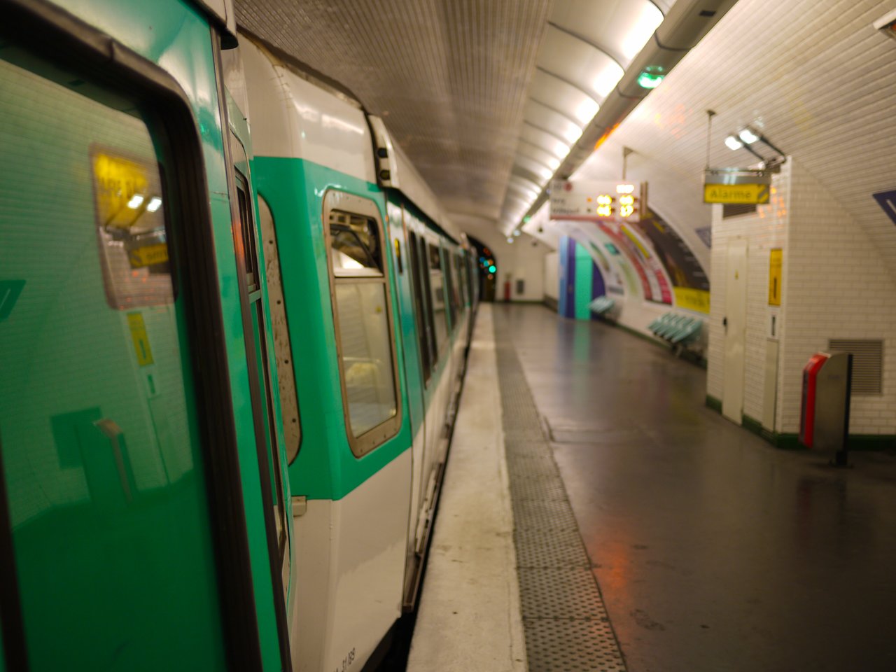 A green and white metro train is stopped at an empty underground station with tiled walls and overhead lights.