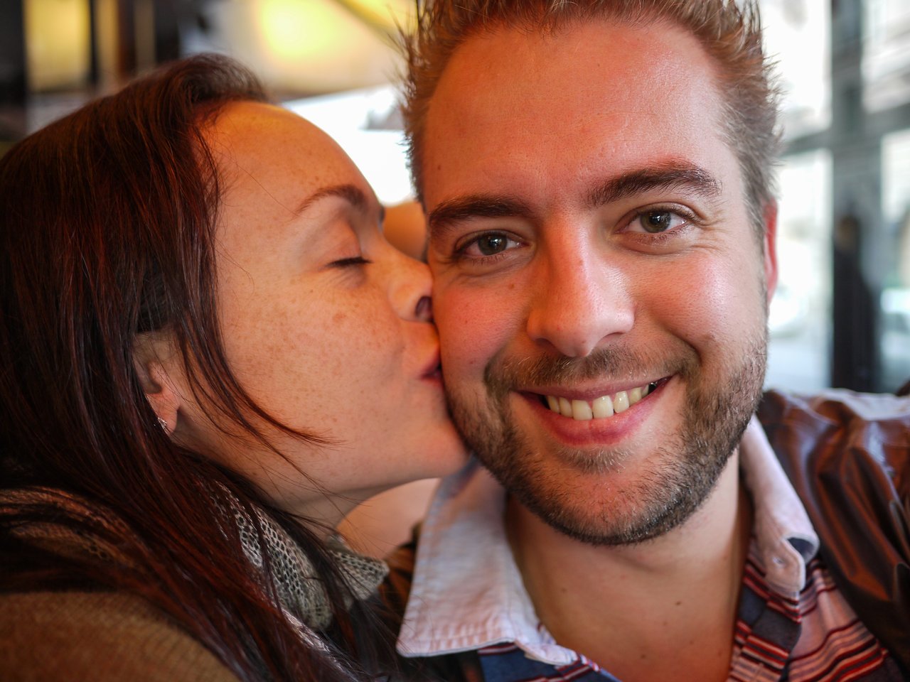 A woman kisses a smiling man on the cheek while sitting together in a café.