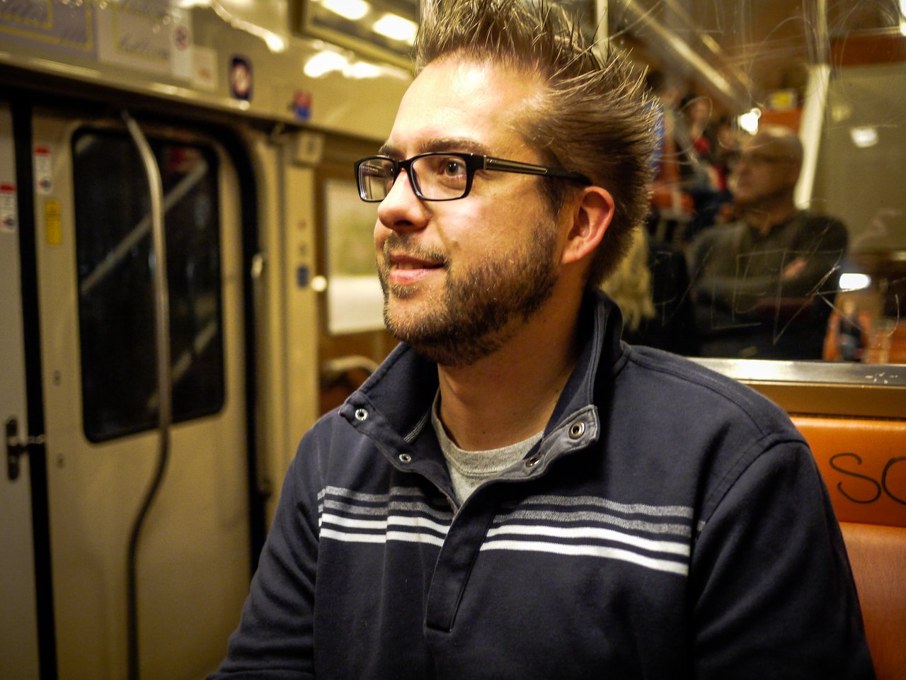 A man with glasses and a beard sits on a train, looking to the side with a slight smile.