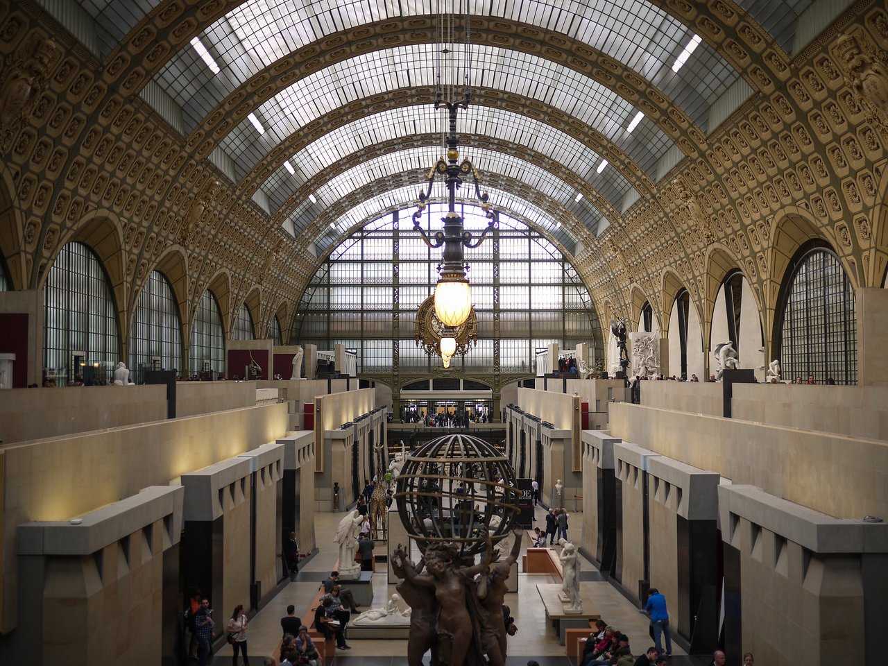 A large museum hall with sculptures, a decorative clock, and visitors walking and observing the artwork.