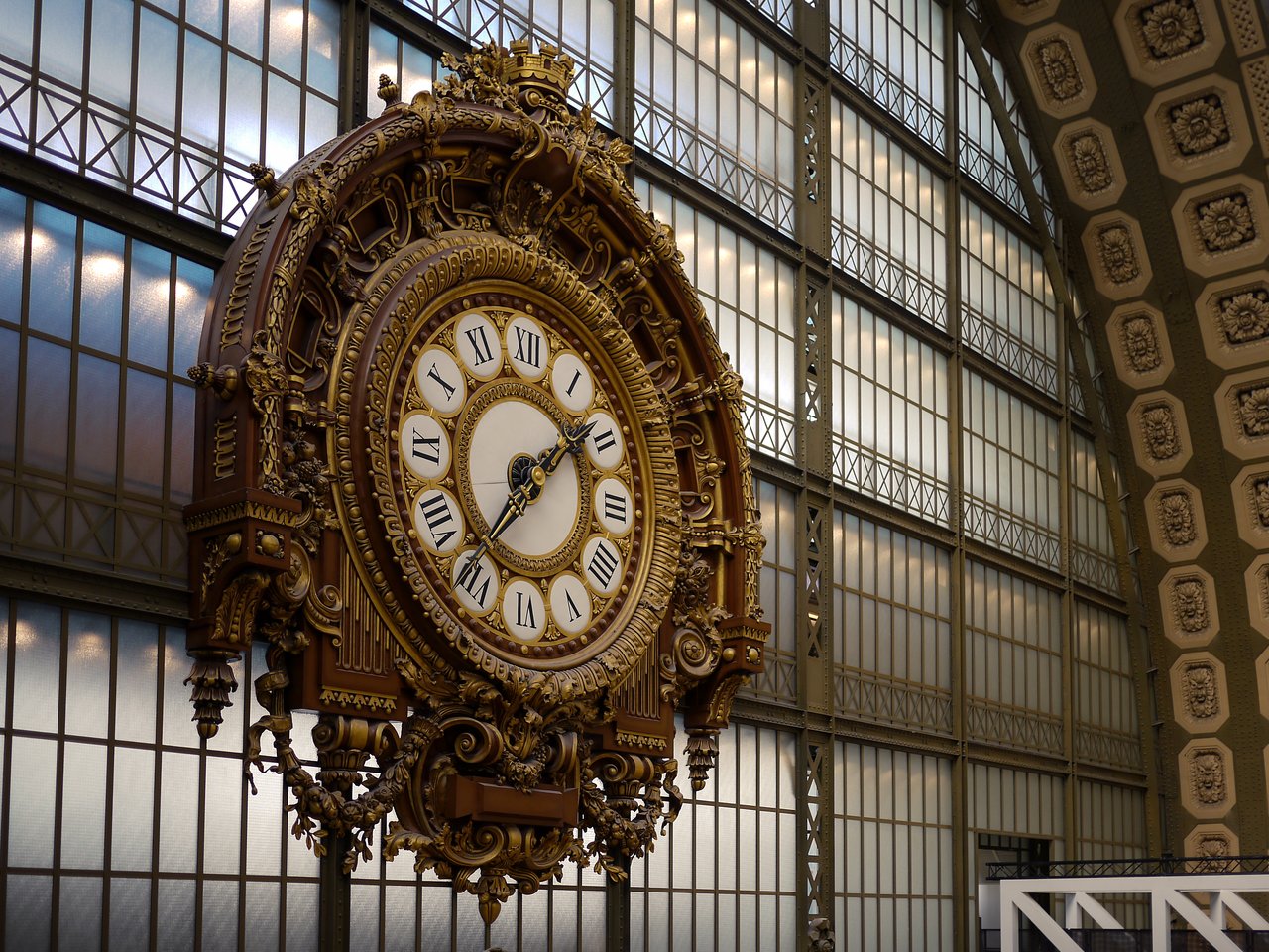 A large, ornate clock with Roman numerals hangs inside a glass-roofed building in Paris.