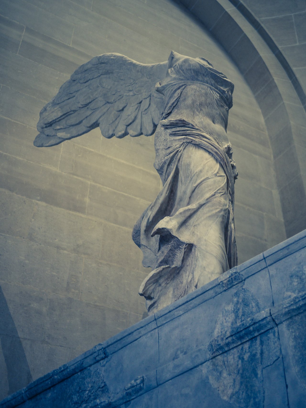 A stone statue of a winged figure without a head, standing on a pedestal against a curved wall.