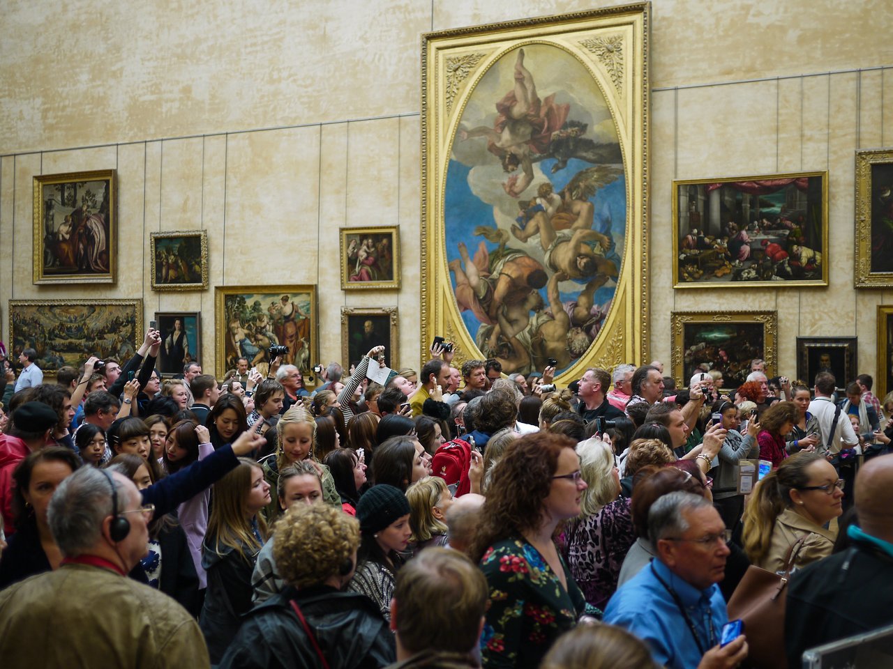 A large crowd of tourists in a museum, many holding cameras and smartphones, looking at an unseen artwork.