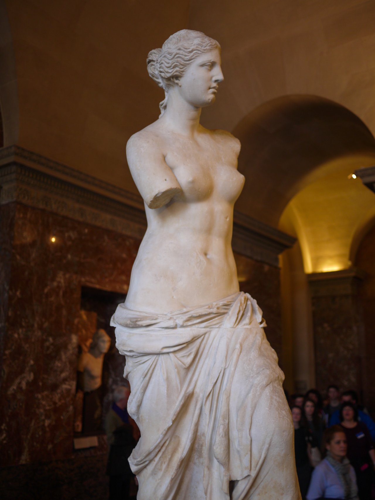 A marble statue of a woman with missing arms stands in a museum, surrounded by visitors.