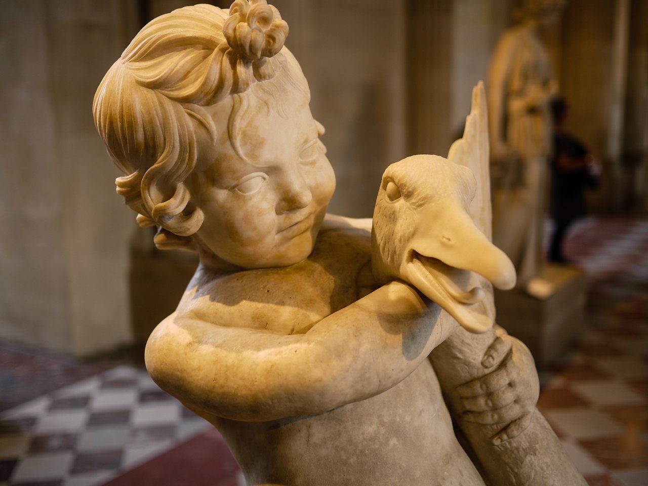 Marble statue of a child holding a bird, with detailed facial expressions and textures, displayed in a museum setting.