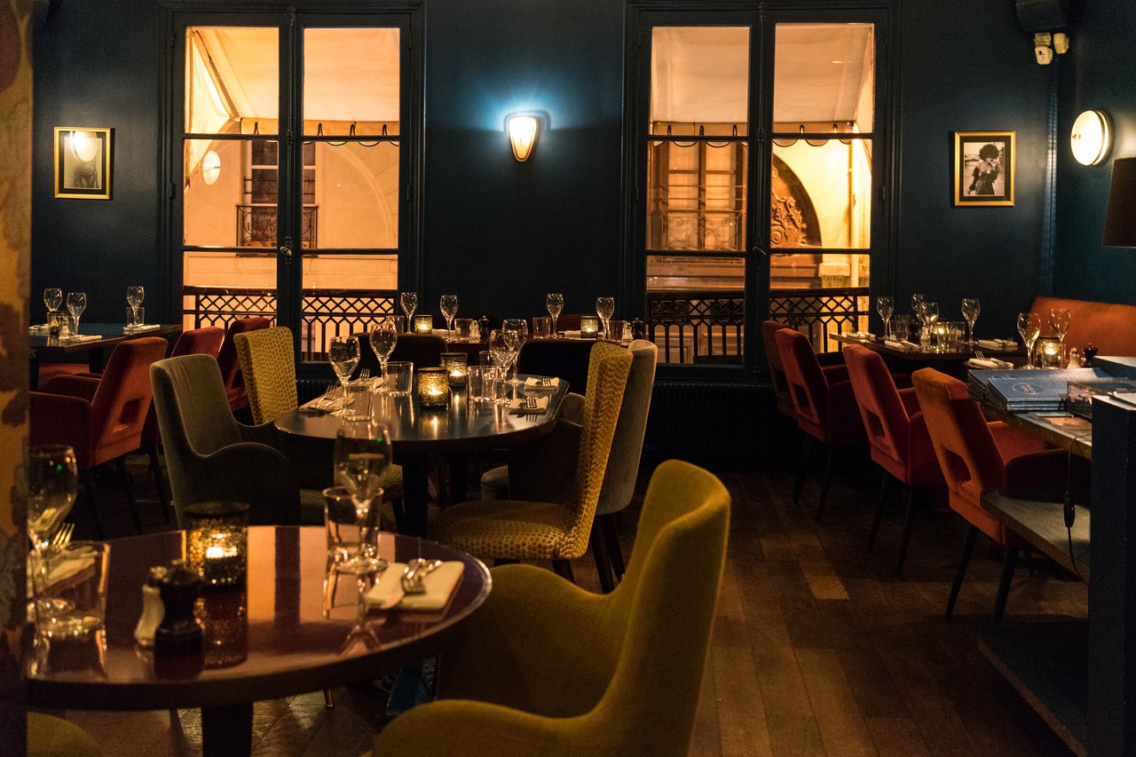 A dimly lit restaurant with set tables, wine glasses, and candles, featuring colorful chairs and large windows.