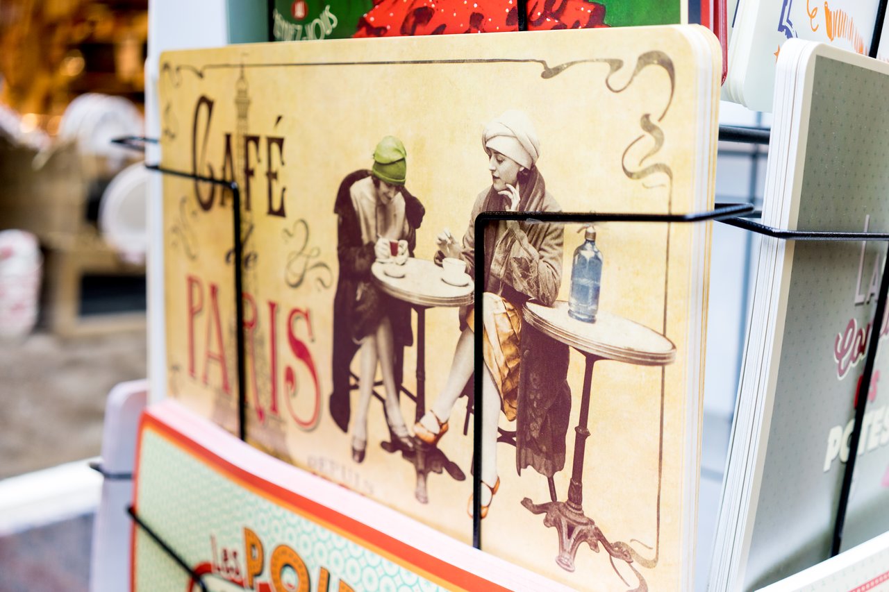 A postcard display featuring a vintage-style "Café de Paris" design with two people sitting at a café table.
