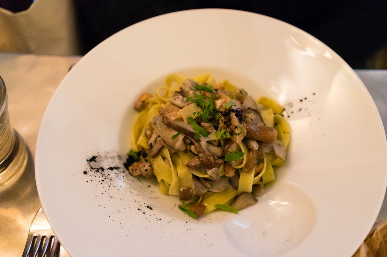 A plate of tagliatelle pasta with mushrooms, garnished with herbs and black seasoning, served on a white dish.