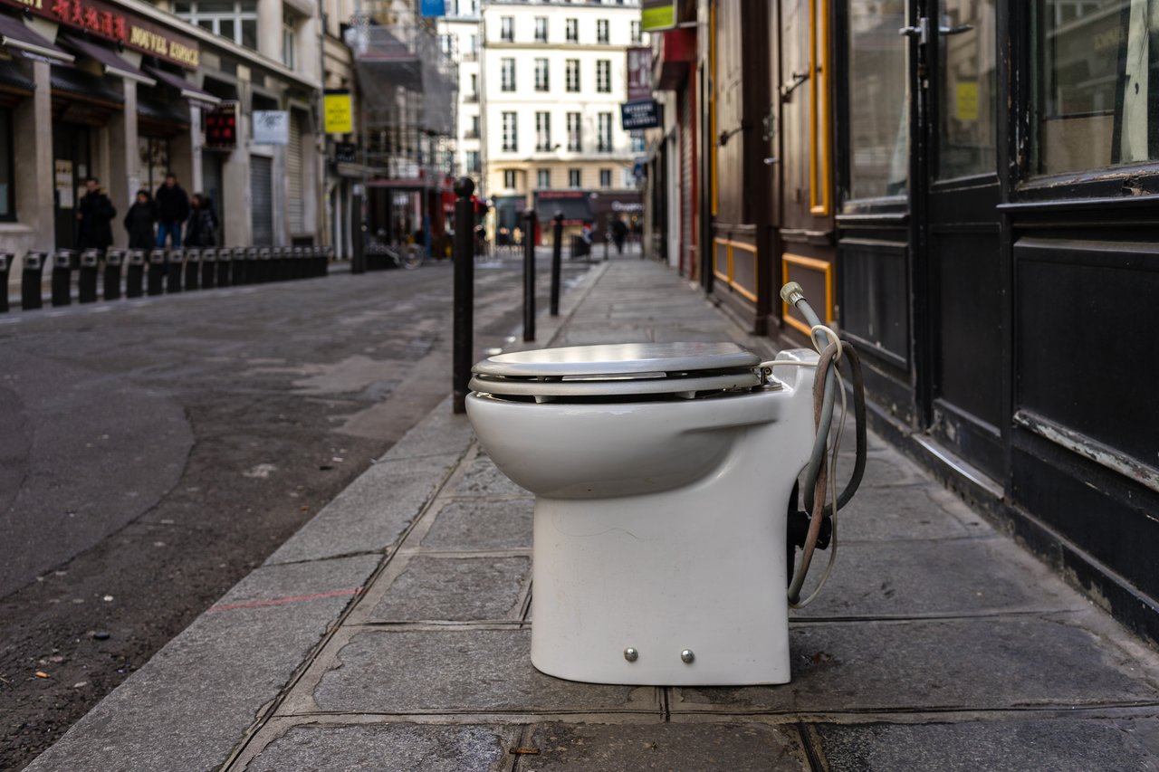 A discarded toilet sits on a sidewalk in an urban street with buildings and pedestrians in the background.
