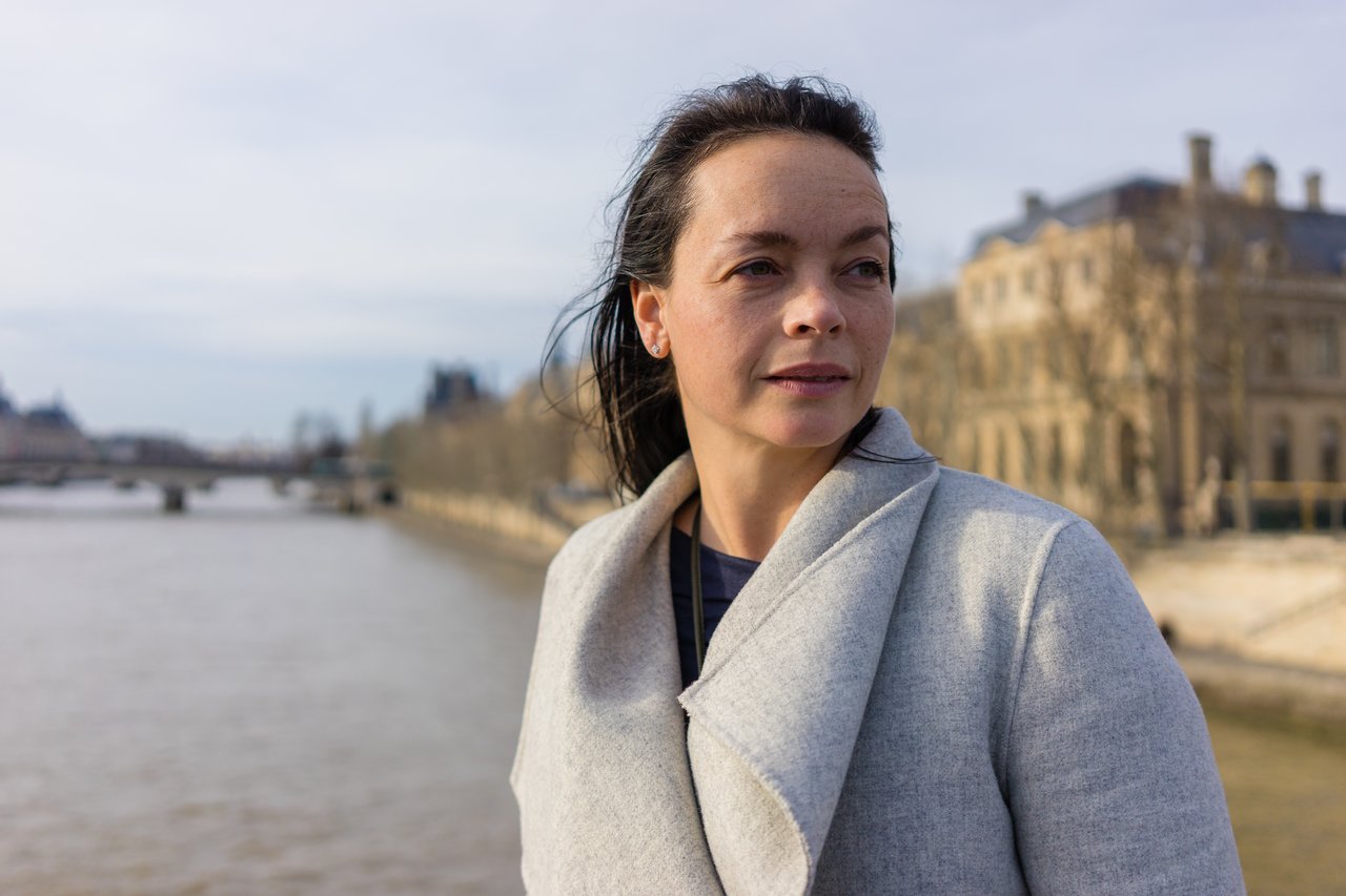 A woman in a gray coat stands near a river, looking into the distance with a calm expression.