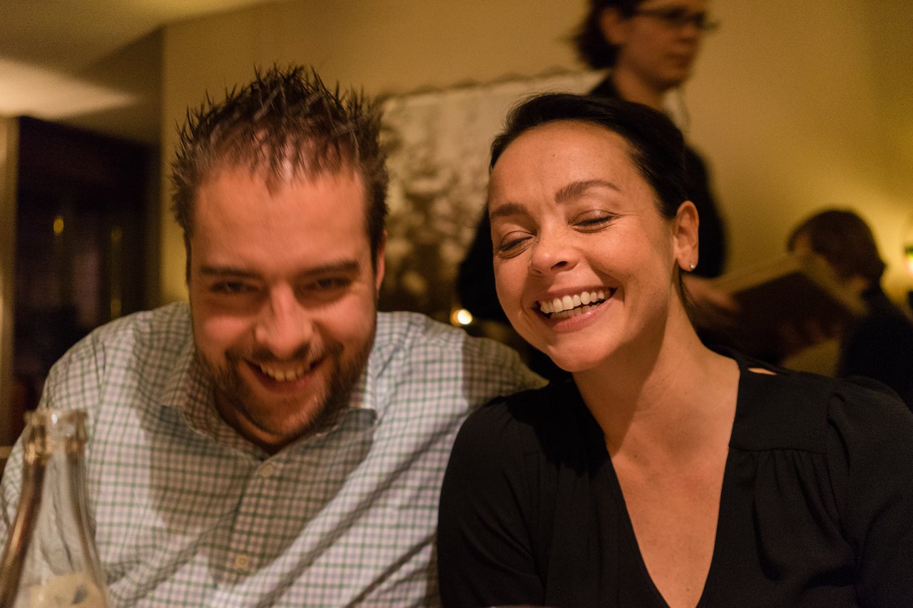 A man and a woman laughing together at a restaurant, with a server in the background.
