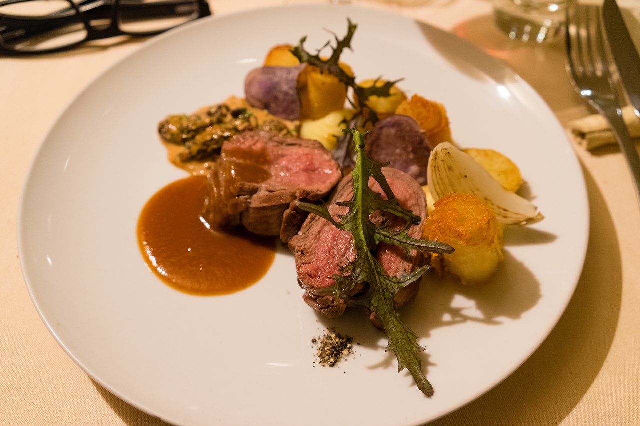 A plated dish with sliced meat, roasted potatoes, sauce, and greens on a white plate in a restaurant setting.