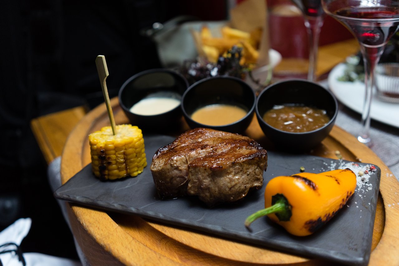 A grilled steak with corn, a roasted pepper, and three dipping sauces on a black plate with wine glasses.