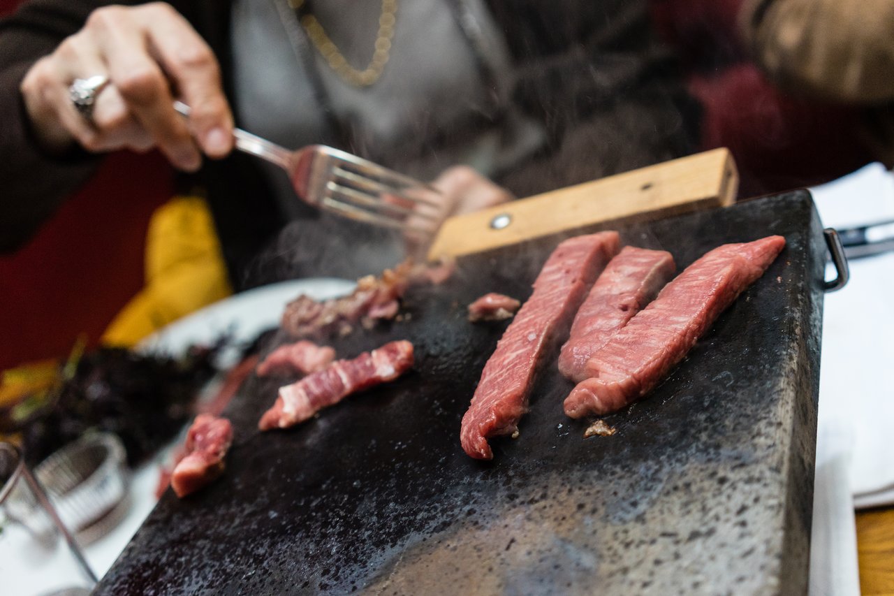 A person cooks strips of raw meat on a hot stone at a restaurant, using a fork to handle them.