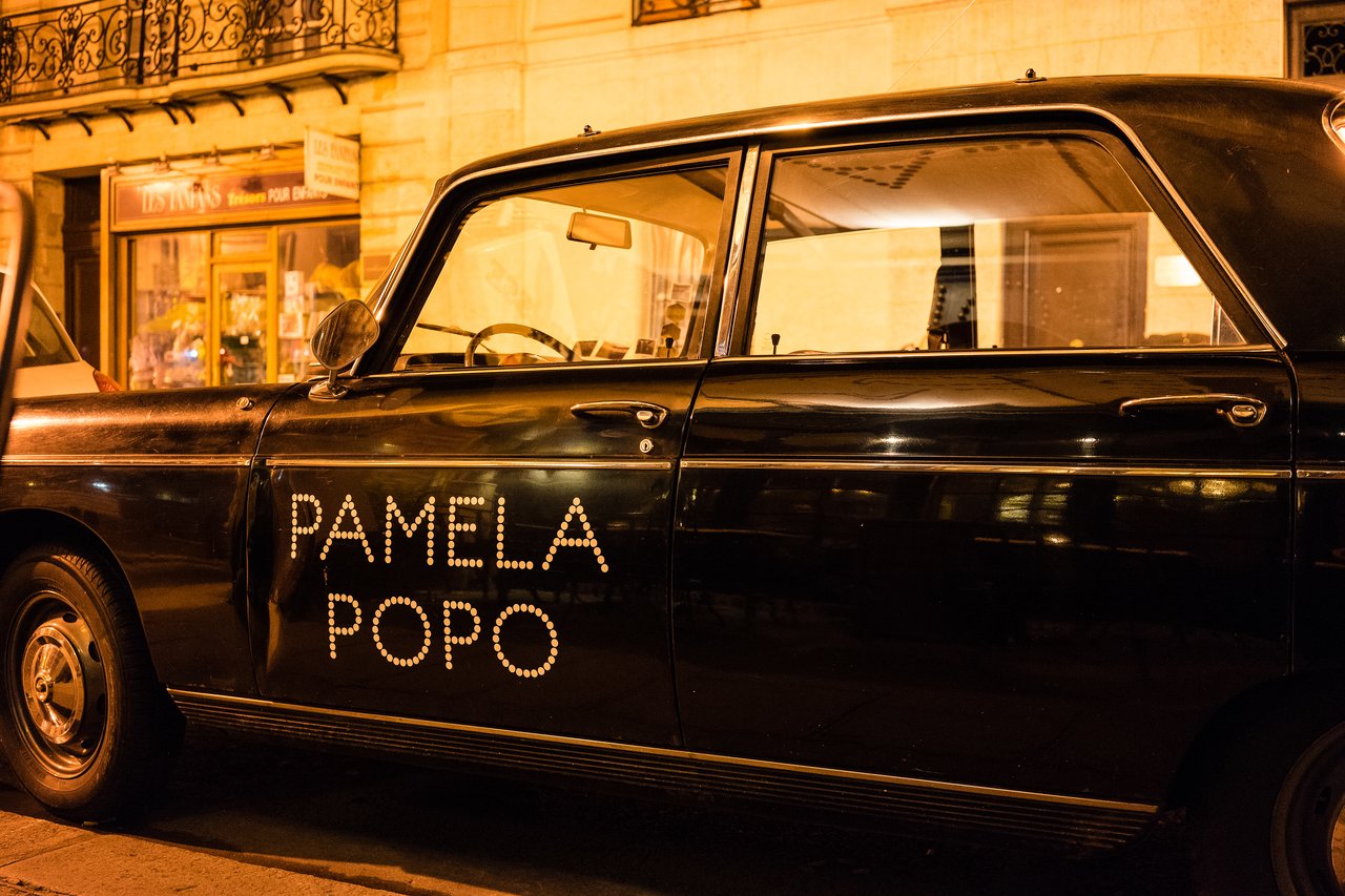 A black vintage car with "PAMELA POPO" written on the door, parked on a city street at night.