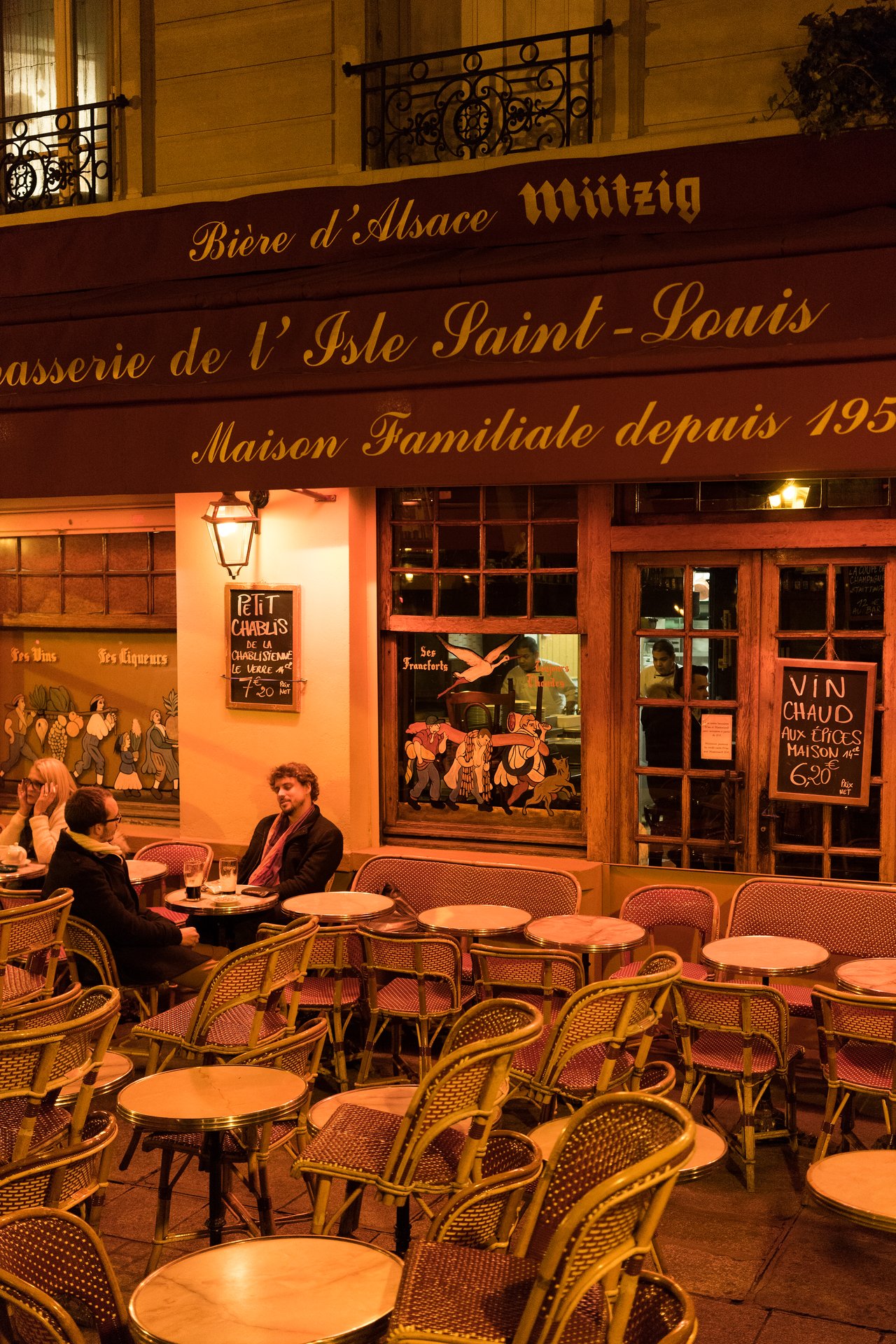 Three people sit at a small table outside a Parisian café, engaged in conversation at night.