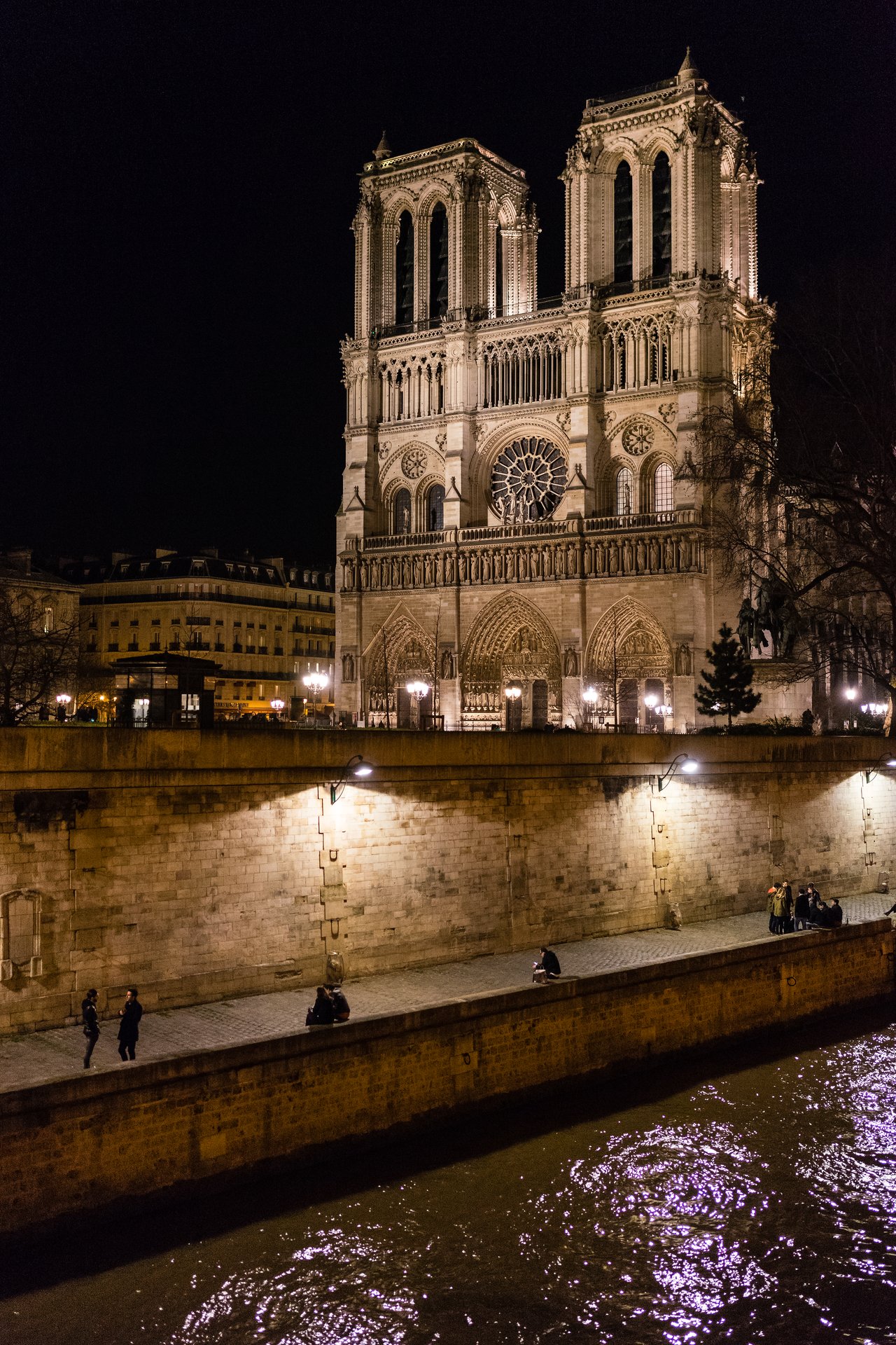 Notre Dame Cathedral illuminated at night, with people walking along the riverbank and reflections on the water.